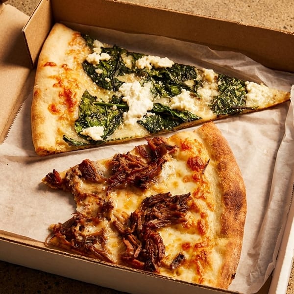 Two pizza slices in a cardboard box, one with spinach and feta, the other with pulled pork.