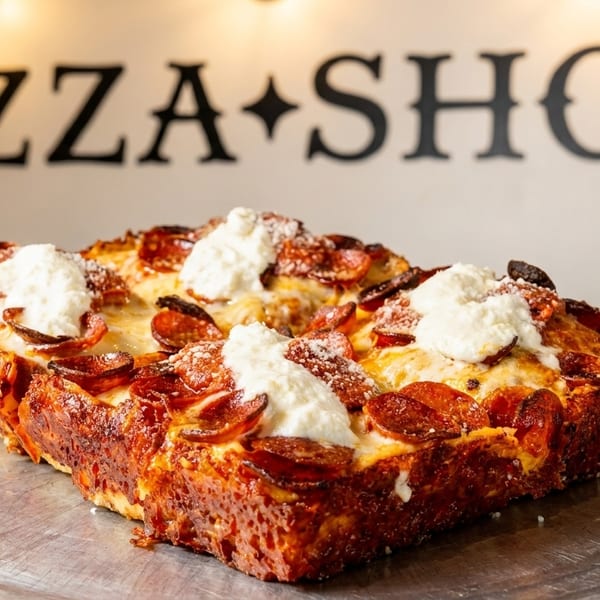 Deep-dish pepperoni pizza with dollops of ricotta cheese on a metal tray, with a "PIZZA SHOP" sign in the background.